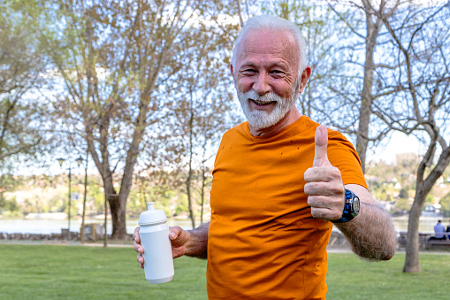 A healthy senior man is catching his breath after exercising in the public city park by the river. Drinking water from a plastic bottle.