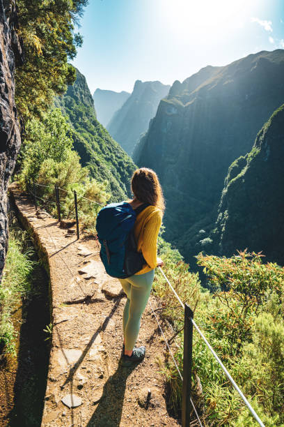 Description: Backpacker woman enjoying scenic view from below large rock wall along water channel at steep cliff through Madeira's rainforest. Levada of Caldeirão Verde, Madeira Island, Portugal, Europe. mujer mochilera disfrutando de la vista panorámica desde debajo de la gran pared de roca a lo largo del canal de agua en el acantilado empinado a través de la selva tropical de madeira. levada de caldeirão verde, isla de madeira, portugal, europa.
