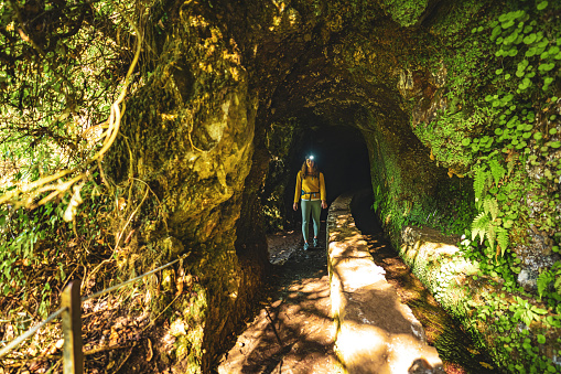 Description: Tourist woman exiting a tunnel along an overgrown jungle trail next to a canal in Madeira rainforest. Levada of Caldeirão Verde, Madeira Island, Portugal, Europe.