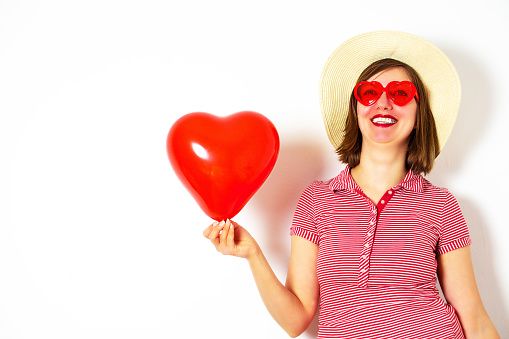 Beautiful smiling woman in red sunglasses and summer hat holding in her hand a red heart balloon for Valentines Day. High quality photo