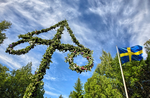 A pole and flag against the blue sky and white clouds. A maypole decorated, covered in flowers and leaves. Pole for celebrating midsummer. holiday. Midsummer traditional Swedish symbol.
