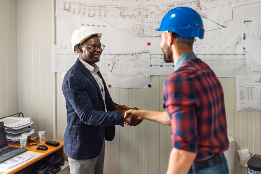 A young African American male civil engineer is shaking hands with his young Caucasian male colleague, who's standing with his back turned.