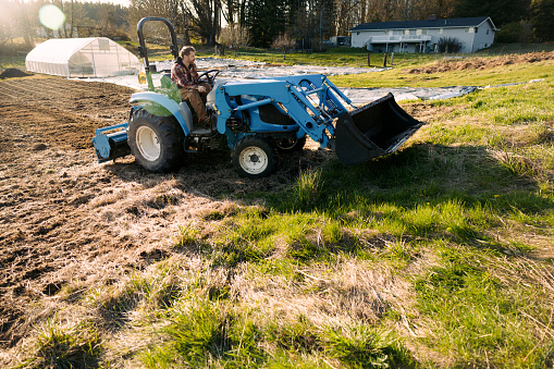 A Caucasian man works on his farm on a beautiful spring day,  using a tractor for the heavy lifting.  He prepares his soil for planting crops.  Shot in Washington state, USA.