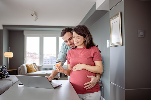 Happy pregnant couple embracing while shopping online via laptop. They are preparing everything for the baby's arrival.