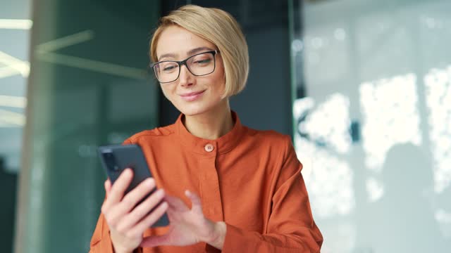 Young female employee wearing glasses is using smartphone while sitting at workplace in modern office.