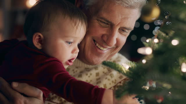 grandfather with baby decorating christmas tree with beautiful ornaments and baubles toddler helping grandpa hang festive decorations at home on calm evening 4k footage