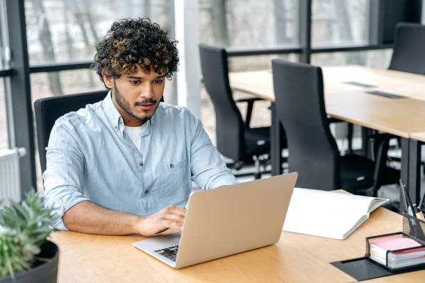 istock Focused smart confident guy of Indian nationality with curly hair, a programmer, IT specialist, sits at a table in the office, works in a laptop, works on a new program, at software support 1483667309