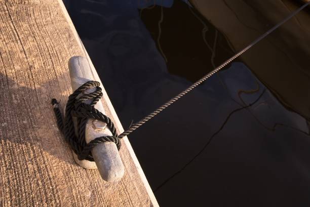 Black braided rope hitched to cleat on dock tied to distant boat seen by reflection in murky waters. Rope illuminated by low afternoon sunlight