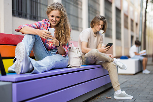 Teenage girl writes message on the smartphone screen while sitting on bench on the street