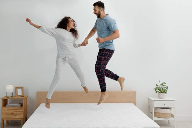 pareja feliz saltando en la cama con colchón cómodo en casa - colchones para habitaciones grandes fotografías e imágenes de stock