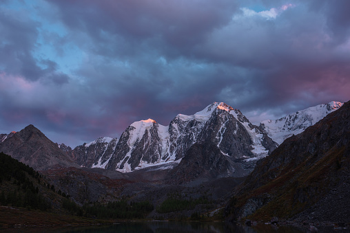 Atmospheric autumn twilight landscape with sunset gold reflection on huge snowy mountain top in violet dramatic sky. Mountain lake with view to giant snow-covered mountains under cloudy sky in dusk.