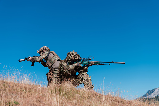 A sniper team squad of soldiers is going undercover. Sniper assistant and team leader walking and aiming in nature with yellow grass and blue sky. Tactical camouflage uniform