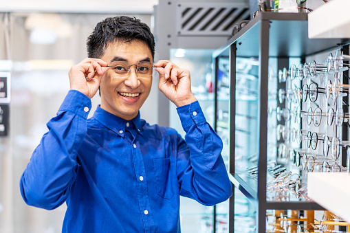Smiling young asian man trying on new eyeglasses in the optical shop. Handsome man wearing new spectacles and looking at camera, Glasses shop, ophthalmology clinic, Choosing glasses concept