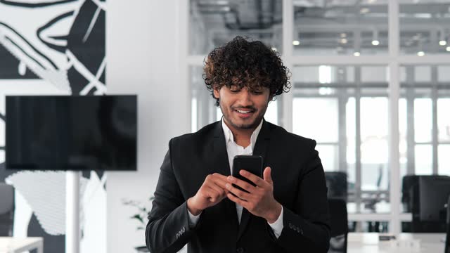 Confident man using mobile phone app in modern office. Indian business professional browsing financial news, smiling reads good news. Successful male staying up to date with latest news and trends.