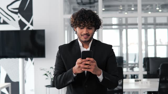 Hispanic man Indian businessman is shown in an office suit, typing a message on his mobile phone, with a happy smile on his face as he engages with social media. Man is working on cell phone.