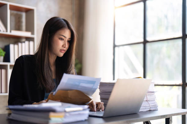 istock Paperwork concept. Asian female manager holding and reading financial documents, Using laptop at workplace in office 1483738737