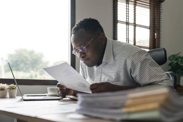 istock Focused serious african american businessman accountant analyst holding documents while using laptop computer screen doing online trade market tech research thinking working sit at home office desk 1483739834