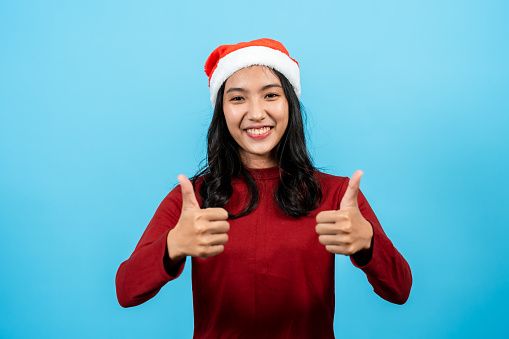 Portrait. Girl with long hair in a red tights wearing a Christmas hat. Raise one hand with a trembling thumb and extend it forward. It's a great gesture. Indoor studio isolated on blue background