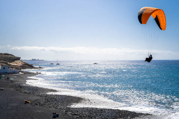 As the sun shines over the Playa de la Enramada beach, a popular paragliding landing zone in Tenerife, an unrecognizable paraglider completing the landing approach over the shimmering Atlantic Ocean.