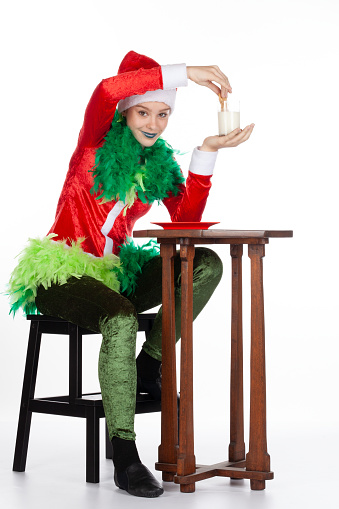 Full portrait of young girl wearing red santa clause hat like grinch perplexed with milk and biscuit, isolated on white background. Human emotion facial expression