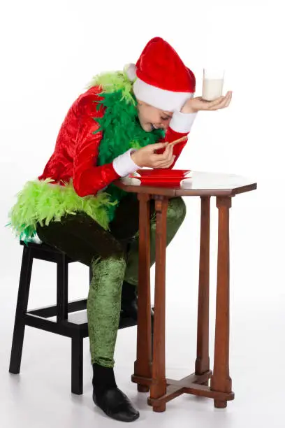 Photo of Full portrait of young girl wearing red santa clause hat like grinch laughing with milk and biscuit, isolated on white background. Human emotion facial expression
