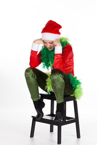 Sitting young girl wearing red santa clause hat like a grinch holding head in hands looking down with smirk, isolated on white background. Negative human emotions