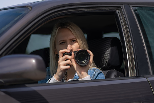 Young blonde woman takes pictures sitting in a car. Concept of journalism, detective, papparation