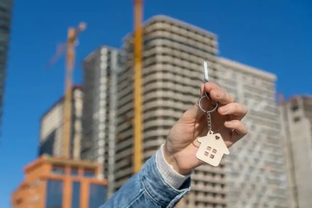 Wooden keychain in the form of a house and a key in a woman's hand against the background of a multi-storey building under construction. Construction, mortgage, rental and purchase of real estate.