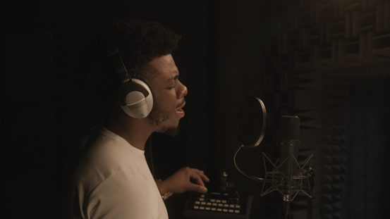 African American vocalist sings lyrical composition into microphone in soundproof room, uses mixing board and gesticulates to audio engineer. Singer works in sound recording studio. Music production.