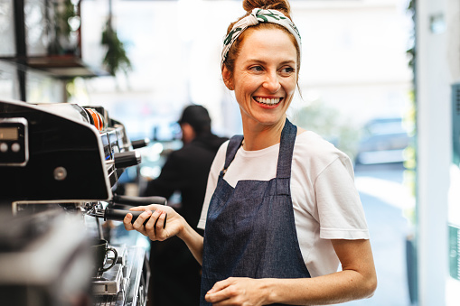 Female coffee shop employee using a coffee machine to prepare coffee for customers. Happy barista using her coffee brewing skills to provide excellent service and hospitality in a cafe.