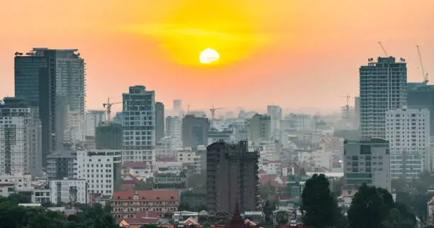 The golden sun hangs low over Cambodia's capital city,it's busy Riverside area on the western banks.Tall modern buildings and apartment blocks loom behind,as sunlight reflects from Tonle Sap river.