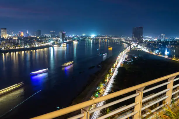 Iluminated buildings and landmarks of Cambodia's capital city,after dusk,streaks of traffic headlights,run along the lively Riverside across the water,street lighting reflecting from the river water.