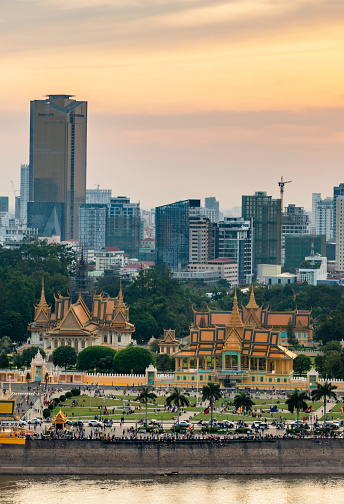 The golden sun hangs low over Cambodia's capital city,it's busy Riverside and famous landmark,the Royal Palace,as sunlight reflects from Tonle Sap river,and tall modern building loom behind.