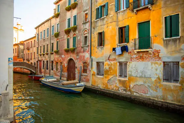 Boat, ancient walls, European architecture, rustic buildings, old city, water canals, waterways. Narrow canal in Venice, Italy sided by buildings with balconies and an old boat in the water.