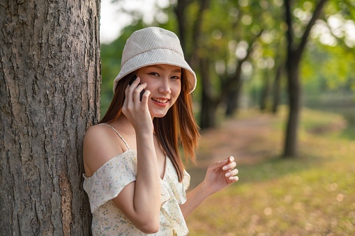 Half body shot of a young asian woman using her mobile phone in a park. Woman smiling happily while talking on the phone.