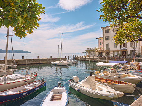 Wide-angle view of the scenic small port of Gargnano, harbouring many different kinds of recreational and fishing boats, and a stretch of its lakeshore promenade, on the western shore of Lake Garda. The bright light of a spring morning, picturesque clouds, leafy branches of citrus trees framing the composition. The outline of the southern extremity of Monte Baldo can be seen on the background. High level of detail, natural rendition, realistic feel. Developed from RAW.