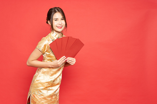 Young Asian woman wearing golden traditional cheongsam qipao dress holding red angpao or packet money gift isolated on red background, Chinese new year concept
