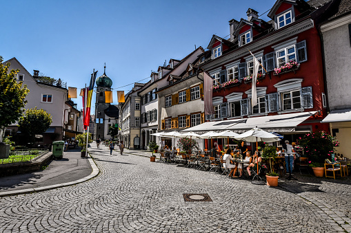 Bregenz, Austria - 16th of August, 2022. Paved Street Along Coffee Bars And Apartment Buildings Ending In Seekapelle Tower.