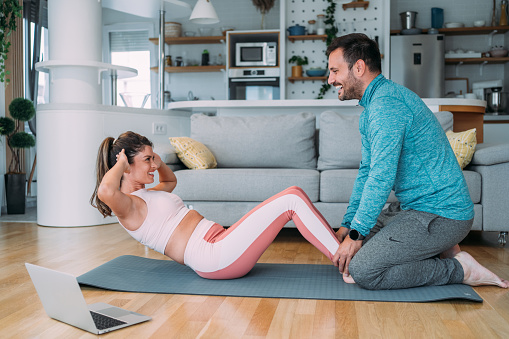 Young couple doing exercises while watching workout video. Young man and young woman in sports wear doing workout at home. Couple using laptop while doing sit-ups. Couple staying in front of laptop and watching online sport lessons. Couple exercising and watching online fitness live streaming classes.