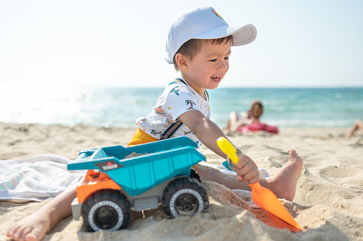 Adorable two year old multiracial baby boy playing with a toy truck on the beach siting on the sand and enjoying the time outside while wearing a hat. Toddler playing with toys in the sand on the beach