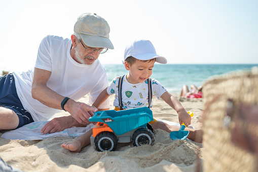 Adorable two year old multiracial baby boy and his grandpa playing with a toy truck on the beach siting on the sand and enjoying the time outside while wearing a hat. Grandfather and toddler grandson playing with toys in the sand on the beach