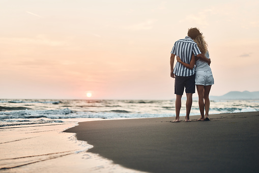 Rear view of embraced couple standing on the beach at sunset. Copy space.