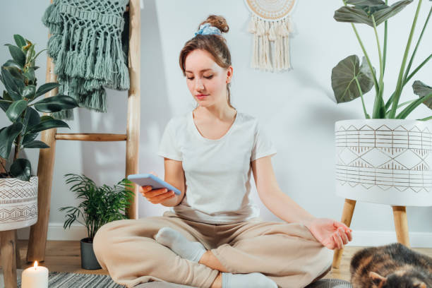 istock Young healthy woman using phone for setting mobile app for meditation at home, sitting in lotus position on floor cushion with relaxed cat pet in modern Scandi interior home. Meditating, relaxed 1483812353