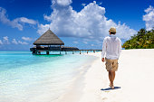 A man in white clothing and sunhat walks down a tropical beach in the Maldives islands