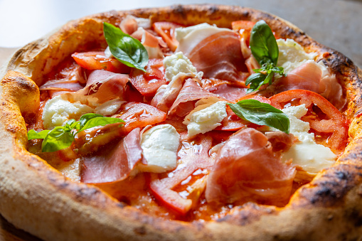 Close-up on round pizza with abundant garnish of prosciutto, mozzarella, tomato slices and basil leaves
