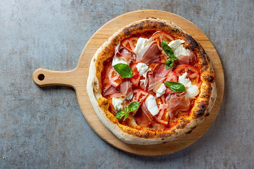 Round pizza with delicious topping, prosciutto, mozzarella, tomato slices and basil leaves, served on wooden board