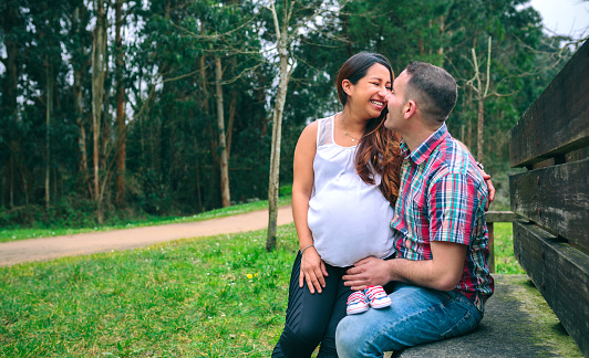 Smiling latin pregnant woman sitting over happy caucasian male with a pair of baby sneakers putted over his legs on nature