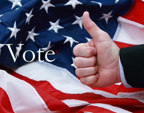 A man giving the thumbs up in front of the American flag with the word vote, Black dress suit