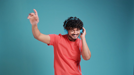Young smiling happy cheerful student Indian man 20s dancing with headphones listen to music have fun in coral t-shirt on blue studio background. People lifestyle concept.
