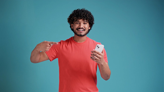 Jubilant exultant happy young bearded Indian man 20s years old hold in hand use mobile cell phone pointing index finger on smartphone, in coral t-shirt on blue studio background.